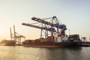 A black container ship stacked with orange containers, sitting below two blue and yellow cranes while docked at a port