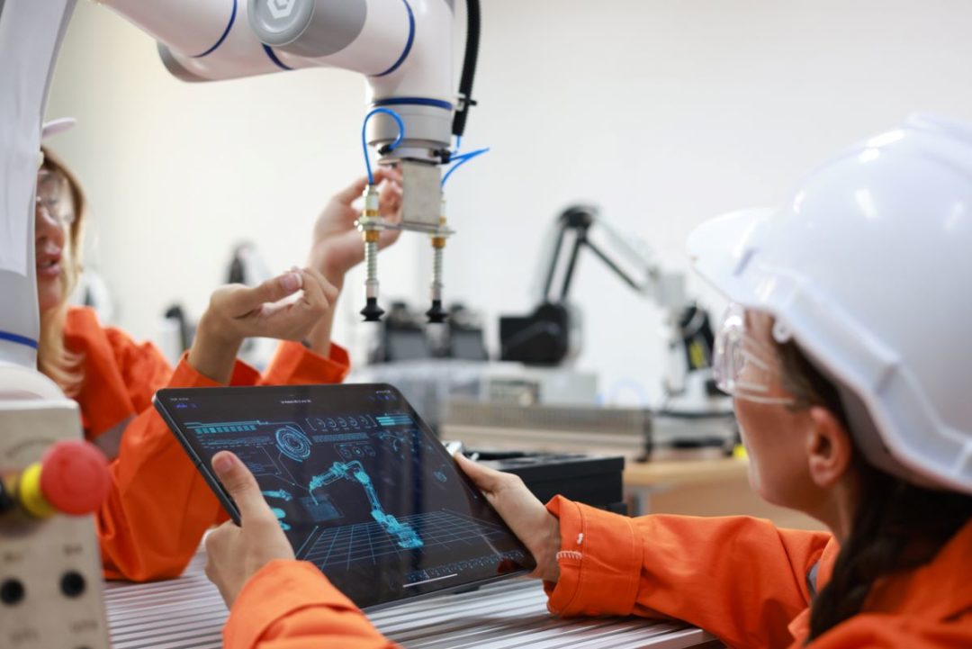 TWO WORKERS IN ORANGE JUMPSUITS AND PROTECTIVE GOGGLES MANIPULATE A ROBOTIC ARM WHILE ONE OBSERVES A COMPUTER TABLET'S SCREEN.jpg