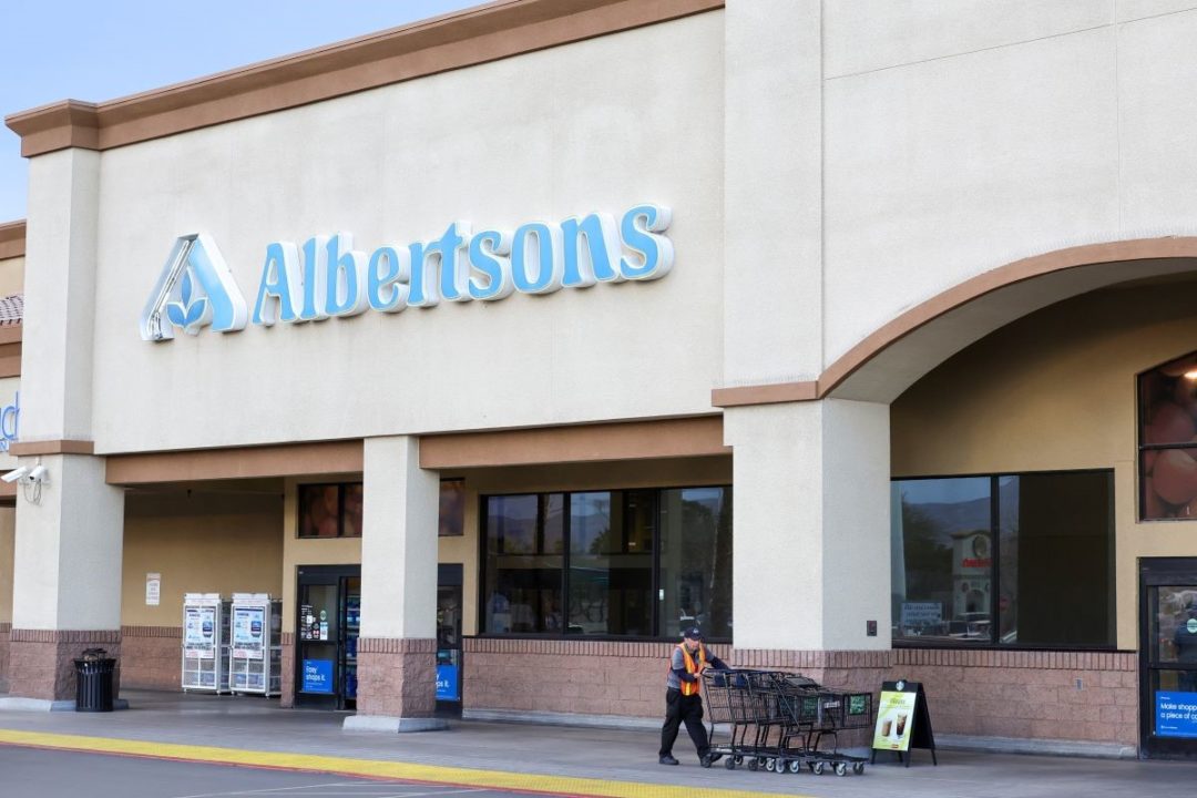 A STORE EMPLOYEE PUSHES SEVERAL LINED UP CARTS IN FRONT OF A GROCERY STORE FRONT BEARING THE NAME ALBERTSONS