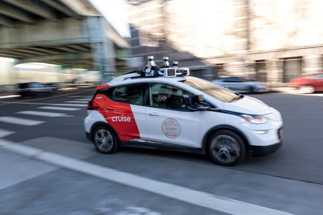 A CAR LABELED CRUISE COVERED IN EXTRA SENSING EQUIPMENT DRIVES DOWN A STREET WITH PEOPLE INSIDE