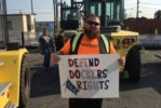 A BEARDED MAN IN SUNGLASSES AND A BRIGHT ORANGE TEESHIRT HOLDS UP A SIGN THAT READS ILA DEFEND DOCKERS RIGHTS, IN FRONT OF A FORKLIFT TRUCK ON A DOCK