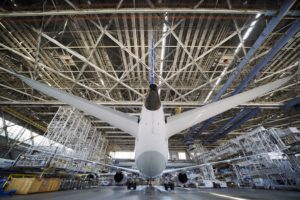 A large white airplane viewed from a low angle, inside of large aircraft hanger with criss-crossed beams across its ceiling