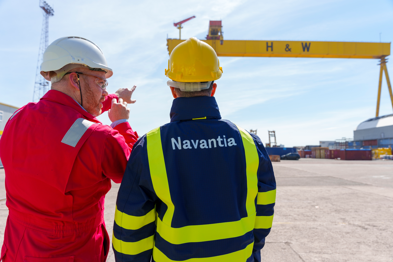 TWO WORKERS IN HI-VIS GEAR ENGAGE IN DISCUSSION UNDER A GANTRY CRANE