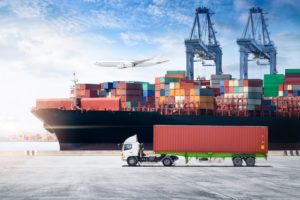 A white truck pulling a red shipping container, with a large black ship stacked with shipping containers that sits below two blue shipping cranes