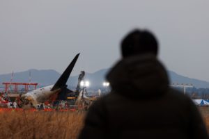 THE WRECKAGE OF A PLANE CRASH IS SEEN IN THE DISTANCE, WITH A SILHOUETTED FIGURE LOOKING ON