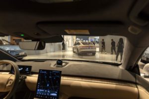 A VIEW OF A CAR SHOWROOM THROUGH THE WINDSHIELD OF A CAR, WITH PEOPLE MILLING ABOUT