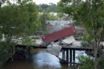 A maroon shipping container tipped over onto another grey container a bridge road, with trees on either side of the image