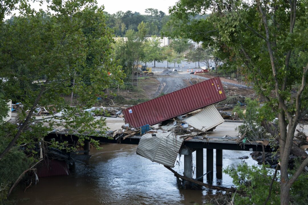 A maroon shipping container tipped over onto another grey container a bridge road, with trees on either side of the image