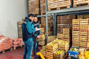 Crates of green and red fruit stacked on metal shelfs, with two men in identical navy blue vests and jeans standing over the crates