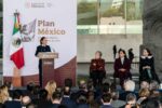 A WOMAN IN A SUIT, HER HAIR TIED BACK, MAKES AN ADDRESS TO AN OUTDOOR AUDIENCE FROM A PODIUM IN FRONT OF THE WORDS PLAN MEXICO AND A MEXICAN FLAG