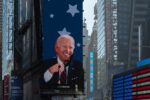 A building in a downtown area with a tall blue sign with an image of Joe Biden smiling, in front of a screen depicting an American flag.