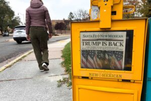 A woman in a purple jacket walking away from a newspaper dispenser, with a paper that reads "TRUMP IS BACK" as the front page headline