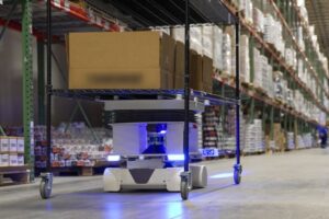 A LIGHTED ROBOT UNIT LIFTS AND CARRIES A WIRE SHELVING UNIT DOWN A WAREHOUSE AISLE