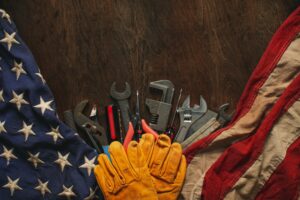 Yellow work gloves set on a brown wood table on top of an American flag, set beneath a row of wrenches and other tools.