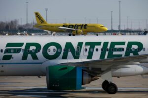 ON AN AIRPORT RUNWAY, A SPIRIT JET PASSES BEHIND A FRONTIER ONE