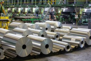 GIANT ROLLS OF METAL SIT STACKED ON A CONCRETE WAREHOUSE FLOOR