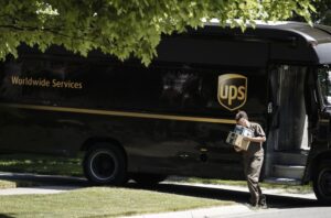 A delivery man in a brown shirt and slacks carrying two small packages up a driveway, with a large brown UPS delivery truck parked on the street behind him