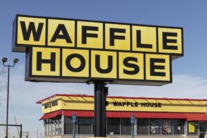 A large yellow sign that reads "WAFFLE HOUSE" outside of a yellow and red diner with the same black lettering across the top. 