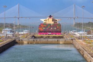 A large container ship with stacks of multi-colored containers and a pink hull moving through a canal, with a grey double-span bridge in the background