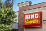 A brick grocery store building with a large red sign over the entrance that reads "KING SOOPERS," with a tree flanking the left side of the frame