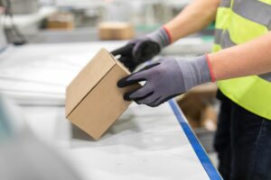 A PERSON WEARING GLOVES AND A HI-VIS VEST INSPECTS A BROWN CARDBOARD BOX ON A CONVEYOR SYSTEM