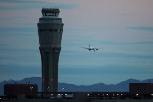 A tall, white conical air traffic control tower, positioned above an airport runway as a plane lands in the distance