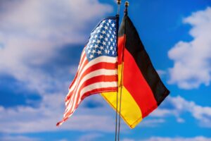 The flags of the United States and Germany positioned next to each other on flag poles, set against a blue sky with sparse clouds