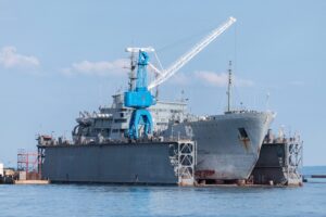 A large gray metal ship docked between two metal walls, with a blue and white crane positioned above the side of the vessel