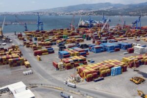 Stacks of multi-colored shipping containers in a concrete lot, with a row of blue cranes along a sea coastline in the background