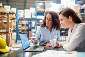 Two women sitting in front of a laptop in a warehouse, inside of a warehouse with rows of metal blue shelving stacked with boxes in the background