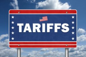 A ROAD SIGN WITH RED STRIPES AND BLUE STARS BEARS THE WORD TARIFFS WITH AN AMERICAN FLAG DECAL