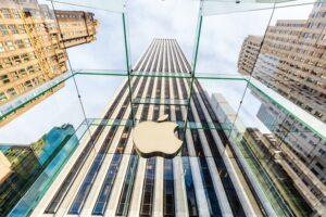 A tall white office building viewed from a low angle, with a large white Apple company logo hanging on a glass overhang over the front of the building