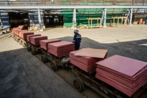 A WORKER IN A HARD HAT SUPERVISES THE MOVE OF MANY SHEETS OF COPPER ON DOLLIES ACROSS A WAREHOUSE DOCK YARD