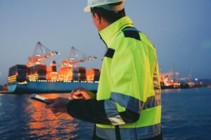 Man in protective gear holding tablet computer in front of harbor terminal at night