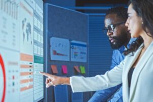 TWO STANDING WORKERS CONSULT TOGETHER AND POINT AT MULTIPLE COMPUTER SCREENS