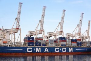 A blue and red striped container ship stacked with shipping containers beneath a row of five white cranes.